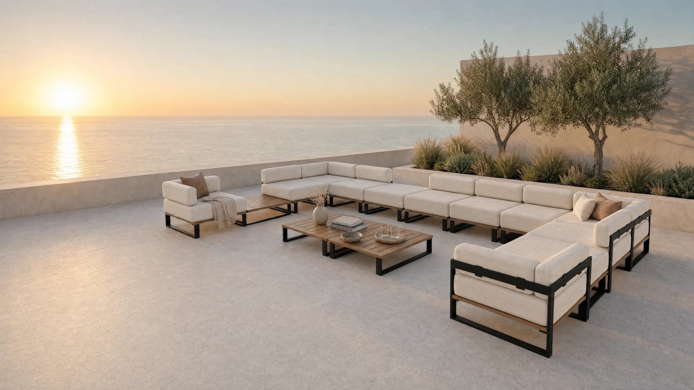 A Nour arrangement on a clifftop terrace at sunset: separate 800 millimetre modules lining three sides of an open centre, each with its own seat cushion and back bolster, two backless poufs breaking the run, and two slatted teak coffee tables standing clear in the middle, with the low sun over the sea beyond.