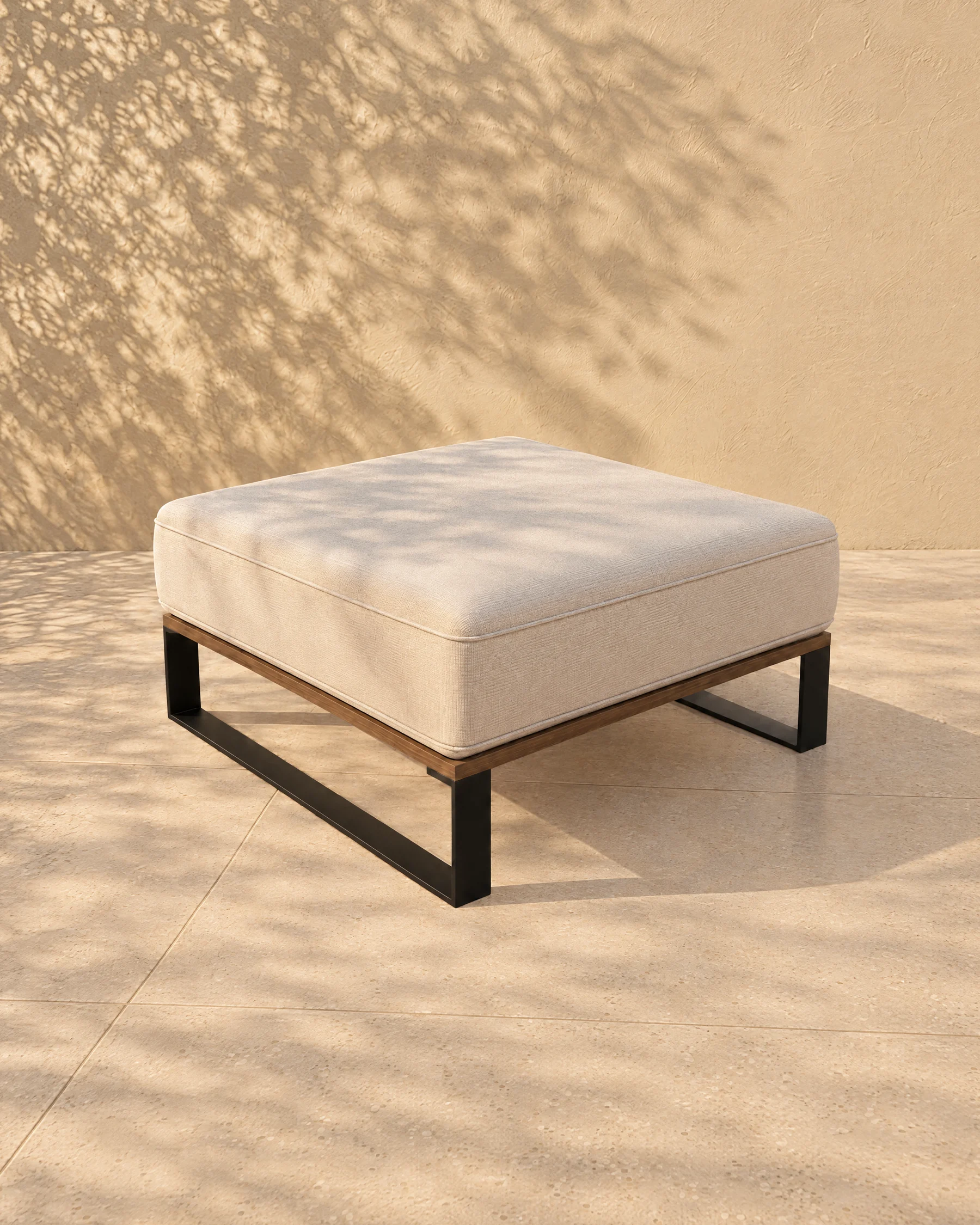 The Nour pouf alone on pale limestone paving before a warm plaster wall: a thin matte black plate sled at each side, a timber deck and one piped seat cushion standing just proud on every side, with nothing above it.